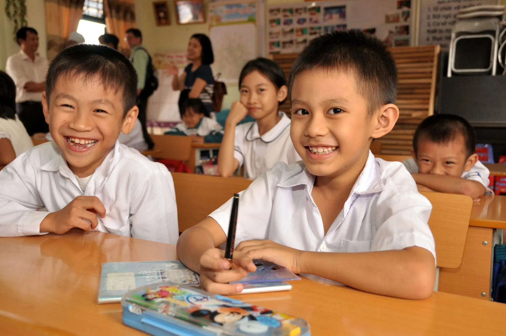 Hai cậu học trò cười tươi bên hộp bút và vở trên bàn học